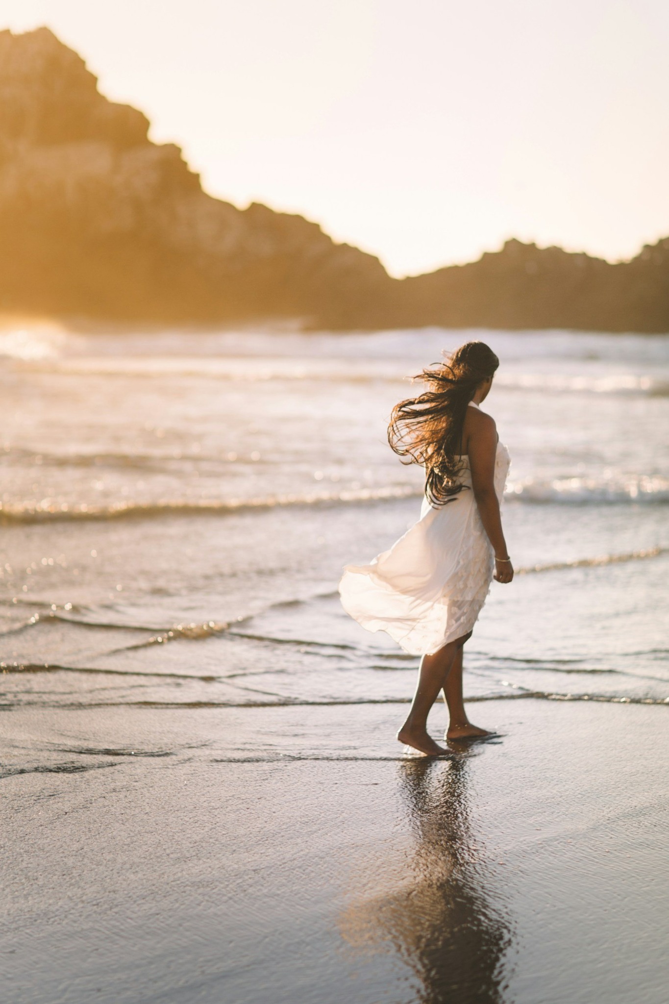 A woman in a cream dress walking beside the sea in warm evening light