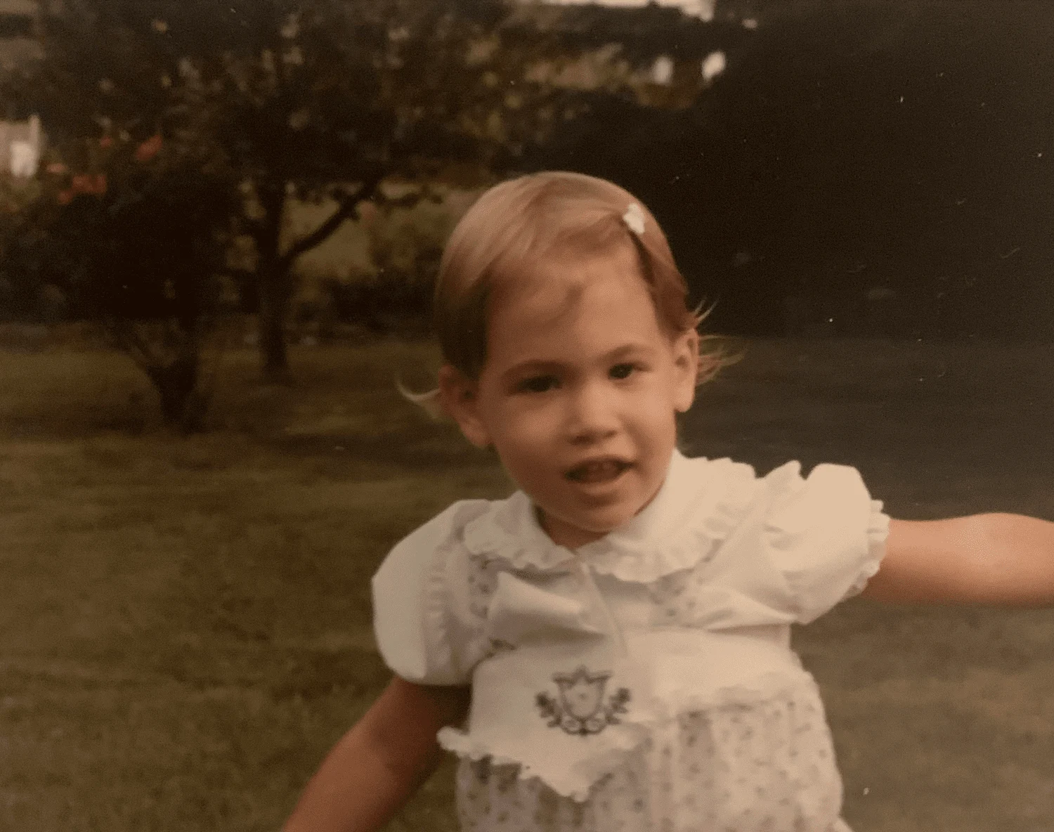 A young girl with short light brown hair, wearing a white dress with lace details and a crest embroidery, standing outdoors in a park at dusk.