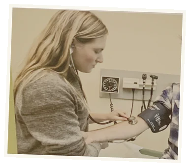 Young woman in a gray sweater taking a person's blood pressure using a sphygmomanometer in a clinical setting.