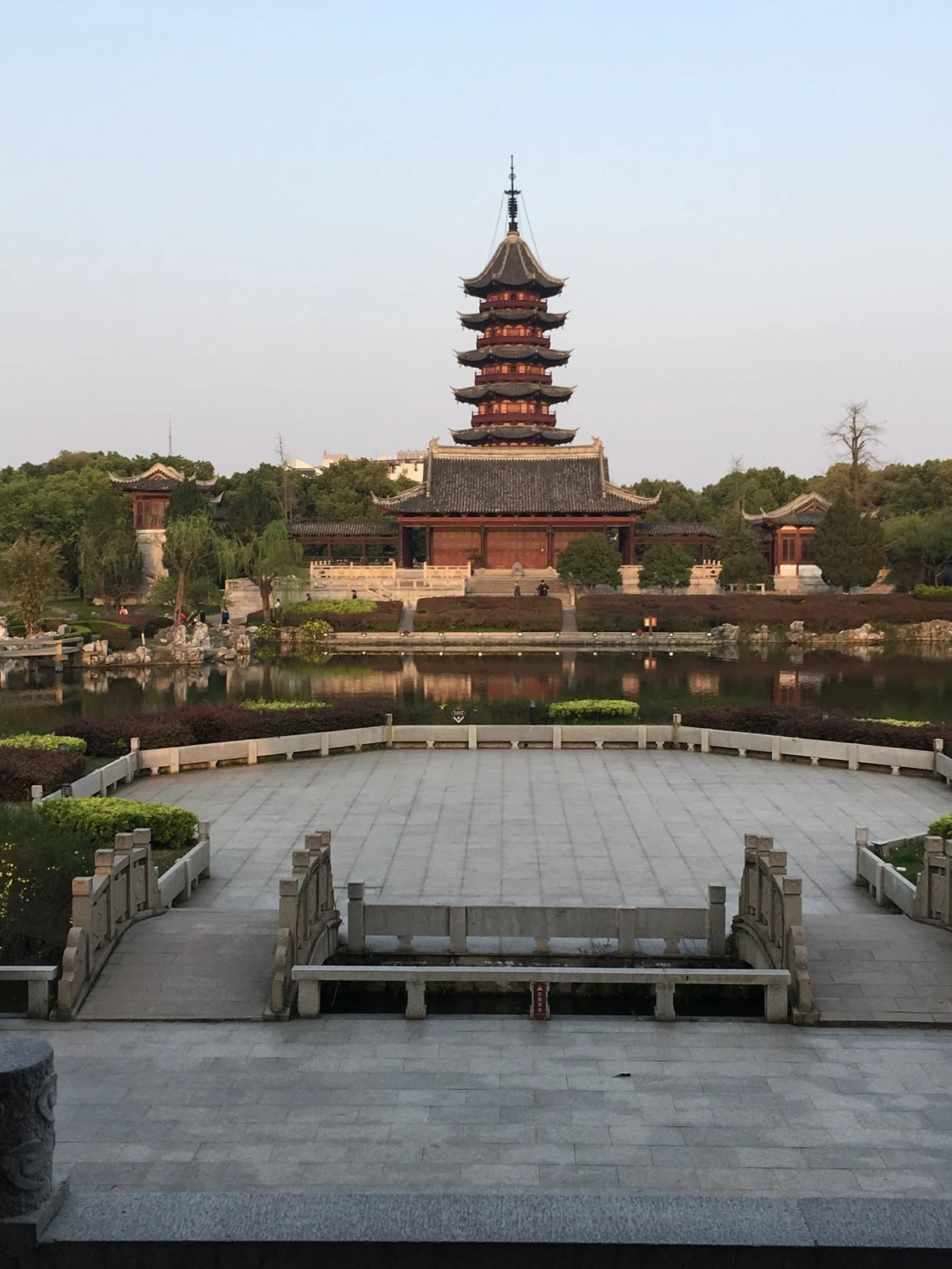 A traditional Chinese pagoda surrounded by trees and a pond with a stone bridge in the foreground.