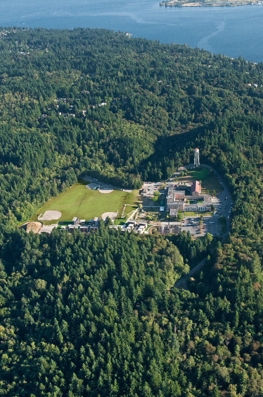 Aerial view of a large green park with sports fields, a parking lot, and buildings surrounded by dense forests near a body of water.